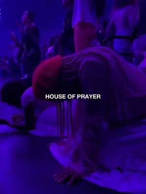 A low-angle shot inside a dimly lit space, likely a church or auditorium, depicts several figures bowing down or kneeling. The primary color is a deep, moody purple, with hints of brighter light sources creating a glow. The focus is on the figures