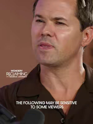 The image shows a close-up of a man speaking, with a blurred background. He appears to be in a studio setting or similar, given the lighting and background color.