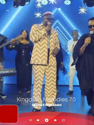 The image shows a group of musicians performing on a stage. The central figure is a man wearing a patterned suit, holding a microphone and singing. Other people, including a woman playing a saxophone or other instrument, are positioned around him. A vibrant blue light illuminates the background, complementing the overall performance atmosphere.