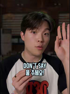 A young Asian man is centered in the frame, looking towards the viewer with a slightly open mouth, as if speaking or about to speak. The background is blurred, possibly a living space, with dim lighting. The man is wearing a black and white baseball-style shirt and a silver ring on his finger. Above him and towards the top of the image is text inside a white box with a neon outline, stating: DON'T SAY 물 주세요!