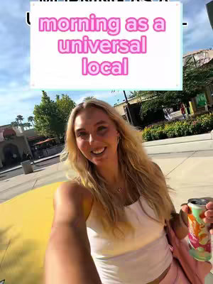 A selfie-style shot with a young woman smiling at the camera. She appears to be at a theme park with buildings and trees in the background. The woman is holding a beverage. Overlayed text appears at the top of the image.