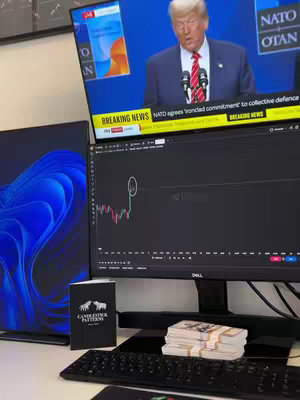 The image shows a workspace setting up with two computer monitors. One monitor displays a news broadcast featuring Donald Trump, while the other displays a stock chart. Resting on the desk is a money stack, a keyboard, and a book labeled "Candlestick Patterns."