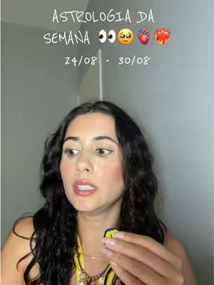 A young woman with dark, curly hair is centered in the frame, looking towards the left with an open mouth. Text over her head says "Astrologia da Semana" in a modern font, with a series of emojis including eyes, a smiling face, a heart, and a flame. The background is a flat, light grey wall.