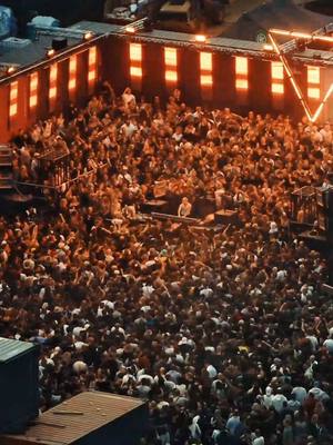 An aerial shot of a large outdoor event, likely a music festival or rave, with a densely packed crowd of people facing a stage area. The scene is illuminated by warm orange and red lights, suggesting late evening or night. The overall composition emphasizes the size of the gathering and the energy of the event.