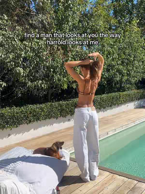A woman with her back to the camera, arms raised behind her head, stands on a wooden deck next to a pool. The woman is wearing white pants and a black top. A small dog lays on a bed in the foreground. Behind her are green bushes and a blue sky. Text overlaid on the image reads, "find a man that looks at you the way harrold looks at me."