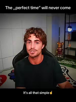 A man with brown curly hair is seated in a gaming chair in front of a background with a shelf and decor. The man is wearing a black t-shirt and is speaking directly to the camera. Text overlays appear at the top and bottom of the image.