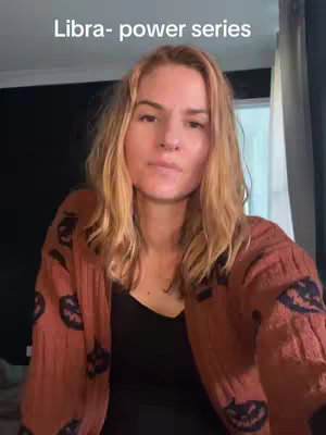 A medium shot features a fair-skinned woman centered, facing the camera. She has shoulder-length blonde hair, and her expression is neutral. She wears a black top underneath a brown cardigan with a pattern featuring pumpkin-like shapes. The background includes a dark wall and the corner of a window with a curtain.