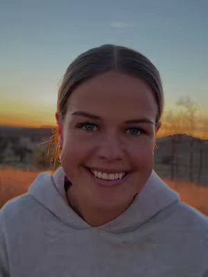 A woman with blonde hair and a gray hoodie smiles broadly at the camera. The background features a vibrant sunset with warm colors.