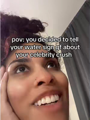 A selfie-style shot featuring a young woman looking upward with a soft smile. Text overlayed on the image reads, 'pov: you decided to tell your water sign gf about your celebrity crush'. The background is blurred, with hints of a room and a dark curtain.