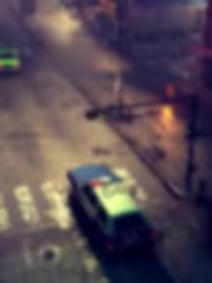 A blurry, top-down shot of a city street at night. A police car with flashing lights is visible on a crosswalk, and another green-colored vehicle is partially seen. The scene is wet, with reflections suggesting recent rain. The overall composition creates a cinematic, atmospheric feel.