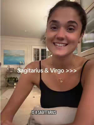 A woman with dark hair smiles at the camera, framed in a home setting. The background includes out-of-focus furnishings and a partially visible window. The text overlay reads "Sagittarius & Virgo >>>" and "IS A SAGITTARIUS".