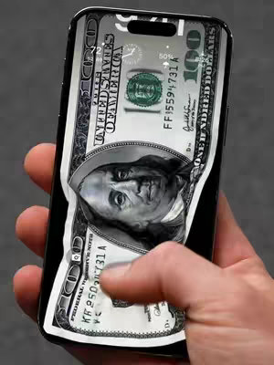 A hand holding a black iPhone with a 100-dollar bill wallpaper on its screen. The bill seems to be peeling off, as if it's part of the background. A gray surface is behind the device.