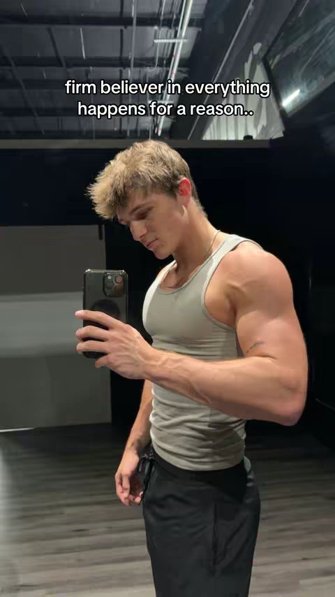 A young, muscular man stands in a gym setting, holding a phone to take a mirror selfie. He's wearing a gray tank top and black shorts, with his arm muscles flexed. A text overlay says "firm believer in everything happens for a reason." The background shows gym equipment and the space has neutral colors.