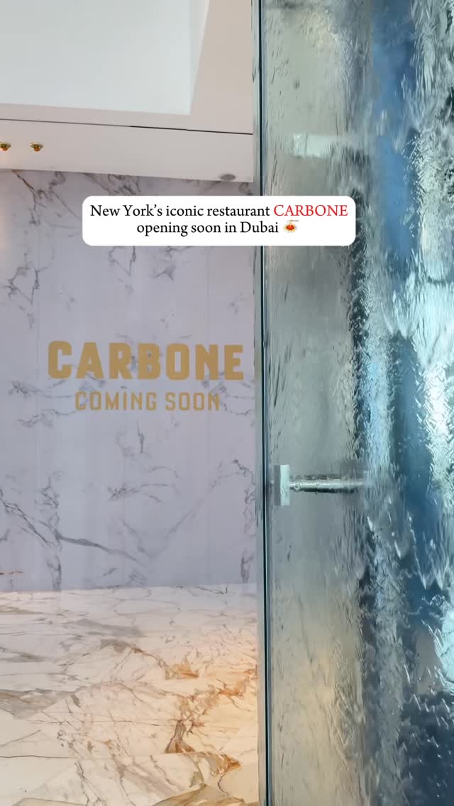 The cover image shows the interior of a high-end establishment. The shot is taken at an angle, focusing on a marble-like textured wall, the restaurant name 'CARBONE' and the words 'COMING SOON' is in the center, along with a glass partition on the right, behind which water is running down like a curtain, and a portion of a ceiling is visible at the top. The overall impression is luxurious and anticipates a new restaurant opening.