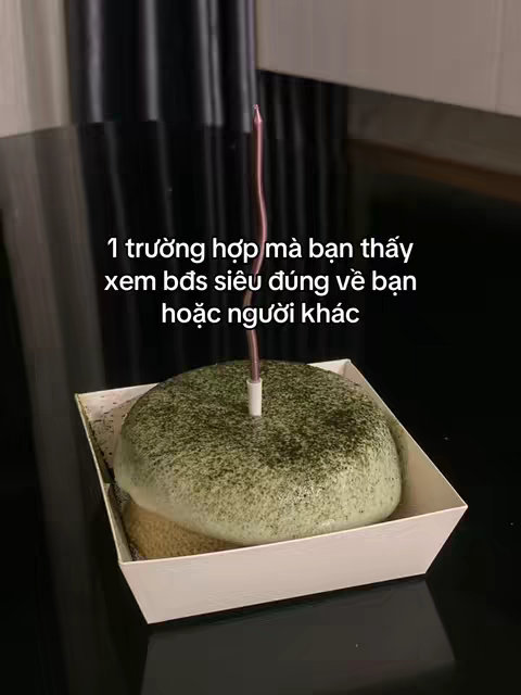 A close-up shot of a dessert resembling a cake with green topping and a candle. It sits inside a white square container on a shiny black surface.