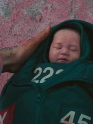 A baby is swaddled in a green tracksuit from the Squid Game series, with the number '22' prominently displayed. A hand with visible veins and red streaks of what appears to be fake blood is holding the baby. The background has a reddish-pink, heavily textured surface. The image is a still from an AI-generated video inspired by the show 'Squid Game.'