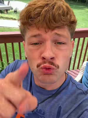 A close-up shot of a young man with light skin featuring a mustache. He's pointing his index finger towards the camera with a slightly puckered expression. He is wearing a blue shirt and a necklace