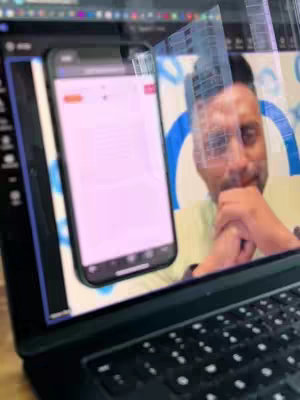A person looks slightly surprised, their face reflected on a laptop screen within a video call interface. On the screen is a phone screen showing a website. The background includes blurry cityscape visuals.