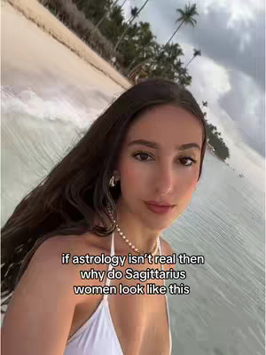 A woman with long, dark hair, in a white top, faces the camera. The background shows a beach with light-colored sand, ocean, and palm trees. Text overlayed on the image poses a question about Sagittarius women.