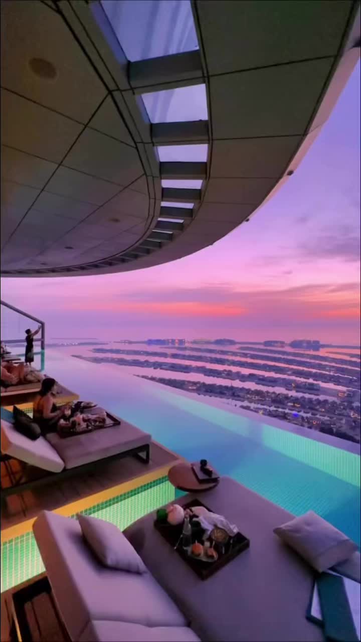 A stunning aerial view of an infinity pool overlooking a cityscape and the sea at sunset, with pink and purple hues in the sky. The pool has people lounging on the side. 