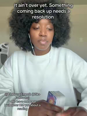 A woman with dark, curly hair and a white sweater is centered in the frame. Text overlays at the top indicate a message, and a user handle and disclaimer appear at the bottom. She holds a deck of cards, suggesting a tarot or psychic reading. The background is a plain wall.