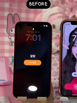 The image shows two iPhones side-by-side, with a pink background featuring a cartoon-like design. The iPhone on the left displays a lock screen with Korean text, time, and an alarm notification. The iPhone on the right shows the creator's face.  The image has text overlayed at the top that reads 'BEFORE' in a black box.