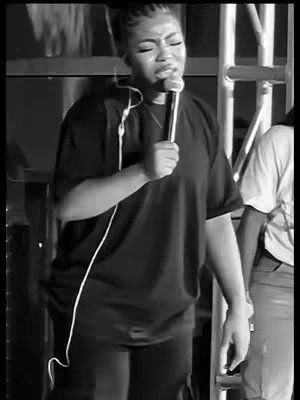 A black and white image of a woman singing into a microphone. The woman is wearing a large, dark shirt, her hair pulled up. She has a microphone in her hand, with the wire going across her body. Behind her is a stage or performance setting. The overall composition and lighting create a scene of a live performance.