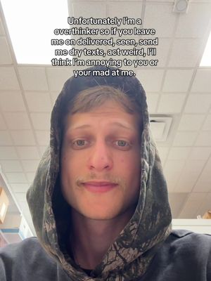 A selfie of a young man wearing a camouflage-patterned hoodie, facing the camera. The image has a light, slightly blurred, indoor background, possibly an office or institutional setting. Text overlay states, 'Unfortunately I'm an overthinker...'