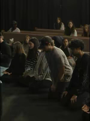 The image shows a group of people in a dim, indoor setting, likely an auditorium or a church. They are kneeling on the floor, facing forward, with some heads bowed. There are rows of wooden benches in the background. The overall scene suggests a moment of prayer or meditation.