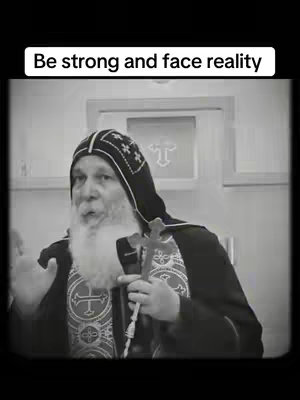 A black and white image of a man holding a microphone and a crucifix. The man is wearing a black hood  with an embroidered pattern and a decorated religious robe. He has a long white beard and is speaking with his hands up. The caption says 'Be strong and face reality' in a white box at the top