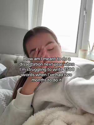 A woman with long brown hair is lying on a bed with a white comforter. She is wearing a white sweater and has one hand up to her face, looking tired or stressed. The background includes a window and a small candle.
