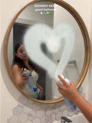 A person is holding a spray bottle and spraying a heart shape on a mirror, with the reflection showing a bathroom setting.