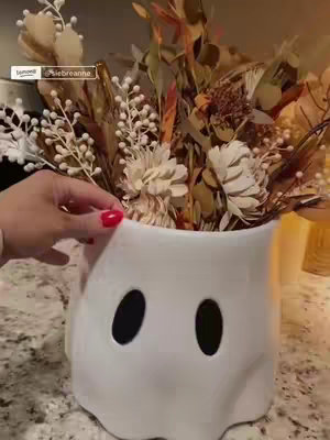A hand holding a white ceramic ghost-shaped vase filled with various dried flowers. The vase has two large oval-shaped cutouts representing eyes. The setting is an indoor kitchen or counter space, with some other decor visible in the blurred background, and a person's hand with red nail polish.