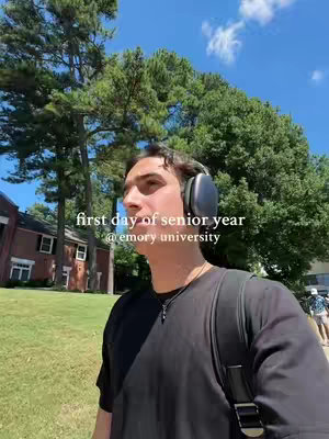 A selfie-style video cover image shows a young man walking outdoors on a sunny day with a blurred background. He has dark hair and is wearing black headphones and a black t-shirt and is carrying a black backpack. The background shows trees, a green lawn, a building, and a bright blue sky. White text is displayed in the center.