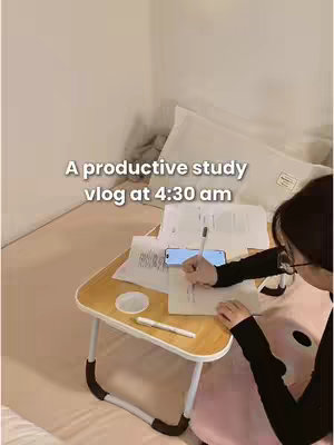 A person is sitting on a bed with a small table in front of them. The table holds papers, a pen, and a phone. Overlay text says, 'A productive study vlog at 4:30 am'.
