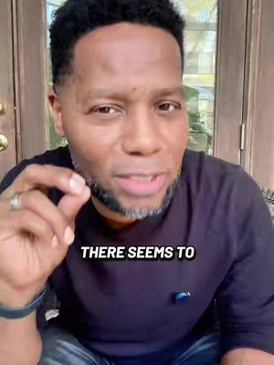 A man with short, dark hair and a salt-and-pepper beard is the primary focus. He is wearing a dark blue crew-neck shirt and is slightly smiling while looking directly into the camera. Overlayed text at the bottom of the image reads 'THERE SEEMS TO'. The background is softly blurred, with a glimpse of a glass door and outdoor scenery.