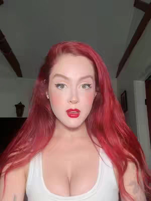 A woman with long, vibrant red hair poses for a selfie. She is wearing a white top and has red lipstick on. The background suggests a well-lit indoor space.