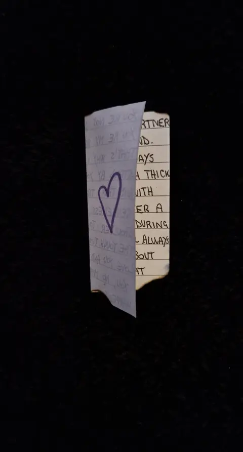 The image shows a piece of paper with handwritten text and a drawn heart, positioned centrally against a black background. The paper is slightly crumpled and has a burnt edge on one side, suggesting it might have been singed or exposed to heat. The text is arranged in lines, and a purple heart has been drawn beneath some of the words. The composition is simple, with the focus entirely on the paper and its message against the dark void.