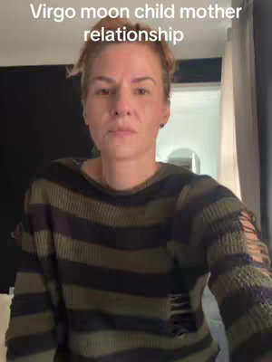 The cover image is a medium shot featuring a woman against a blurred background with text overlayed, reading "Virgo moon child mother relationship". The woman, who appears to be in her mid-30s to late 40s, has light skin, fair brown hair and a focused expression. She wears a dark green and black striped sweater with distressed areas. The background is a dimly lit room, with a dark wall and a glimpse of a curtain to the right. The lighting is soft, illuminating the woman's face, with the text overlayed at the image's top.