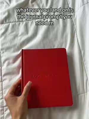 A hand holding a red journal lies atop a wrinkled white bedsheet. Above the journal, overlaid text reads 'whatever you land on is the journal prompt you need rn'.