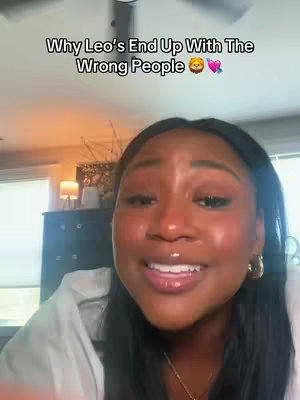 A selfie-style video cover image featuring a woman with brown skin, angled slightly to the right, smiling. The background is softly lit with a lamp visible to the left. Text overlayed at the top reads 'Why Leo's End Up With The Wrong People'