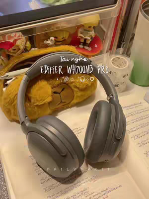 The cover image showcases a pair of Edifier WH700NB PRO headphones placed on open notebook pages. The headphones are a dark gray color, positioned prominently towards the center. In the background, there's a partially visible clear container filled with small toys, a fluffy yellow lion-shaped pencil case, a white ceramic tumbler, and handwritten notes, suggesting a study or workspace environment. The lighting appears soft and natural, with a warm overall tone.