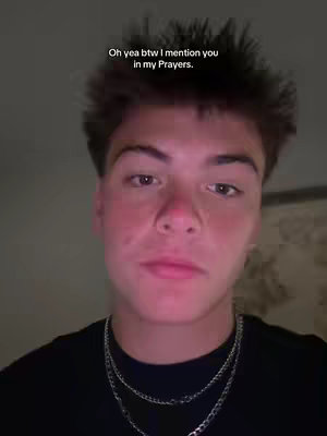 A selfie-style shot of a young man. He has short, dark hair and is positioned in front of what appears to be a plain wall or interior setting. The lighting is soft and appears to be ambient . He is wearing a black shirt and a chain necklace. The image is overall a medium shot, focusing from the top of the chest to the top of head.