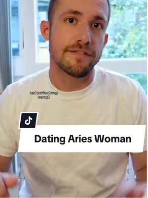 A man in a white t-shirt is facing the camera, the background appears to be an interior with a window. A text overlay reads "Dating Aries Woman".