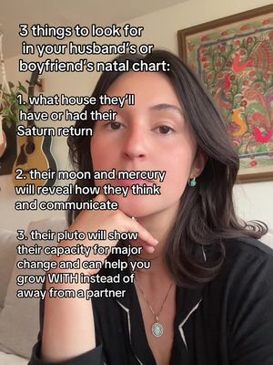 A selfie-style shot with a woman in the foreground. She is looking at the camera with a neutral expression, her hand gently touching her chin. Behind her is a warm-toned room with a painting and a few decorative items. Text overlays detail astrological insights.