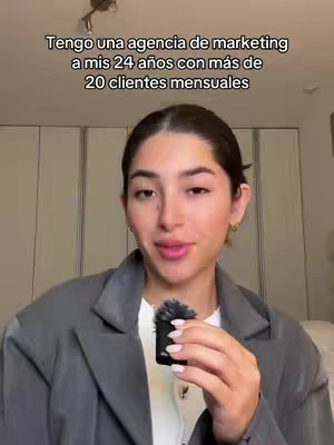 A woman with dark hair in a grey blazer is holding a small microphone. The background is a blurred interior with neutral tones. Text overlay reads: 'Tengo una agencia de marketing a mis 24 años con más de 20 clientes mensuales'.