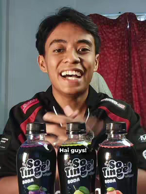 A young man with dark hair smiles broadly, holding three bottles of So Tinge soda. The bottles, featuring colorful liquid in shades of purple, green, and red-orange, are positioned in the foreground. Behind him is a blurred background with a red curtain and a neutral wall, creating a simple setting.