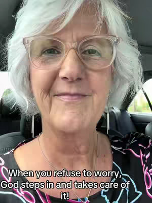 A mature woman with short, wavy gray hair and glasses is seen in close-up, smiling gently. She is wearing a dark top, and in the background is the interior of a car.