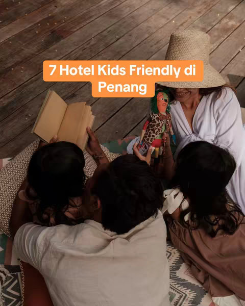 An overhead shot shows a group of people gathered on a wooden surface, possibly a deck, with a colorful patterned blanket or mat spread out beneath them. One person is holding an open book, appearing to read to two children who are leaning in to listen. A fourth person, possibly an adult, is holding a doll. A warm orange text overlay reading '7 Hotel Kids Friendly di Penang' pops against a light background.