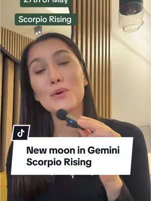 The image shows a woman with dark hair and makeup, holding a microphone and speaking. There is a text overlay at the bottom that says 'New moon in Gemini Scorpio Rising.' Behind her is a light-colored wooden wall.